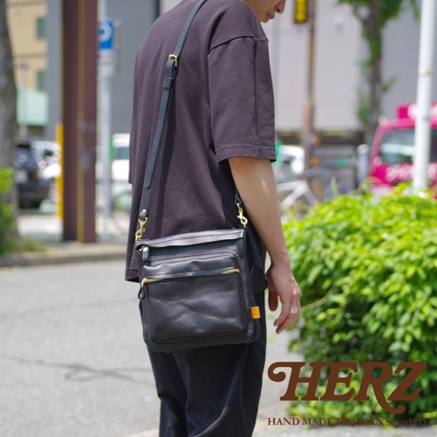 黑色皮革製作的直式斜背包，配有金色拉鍊及可調式肩帶，包身前方有兩個拉鍊口袋。