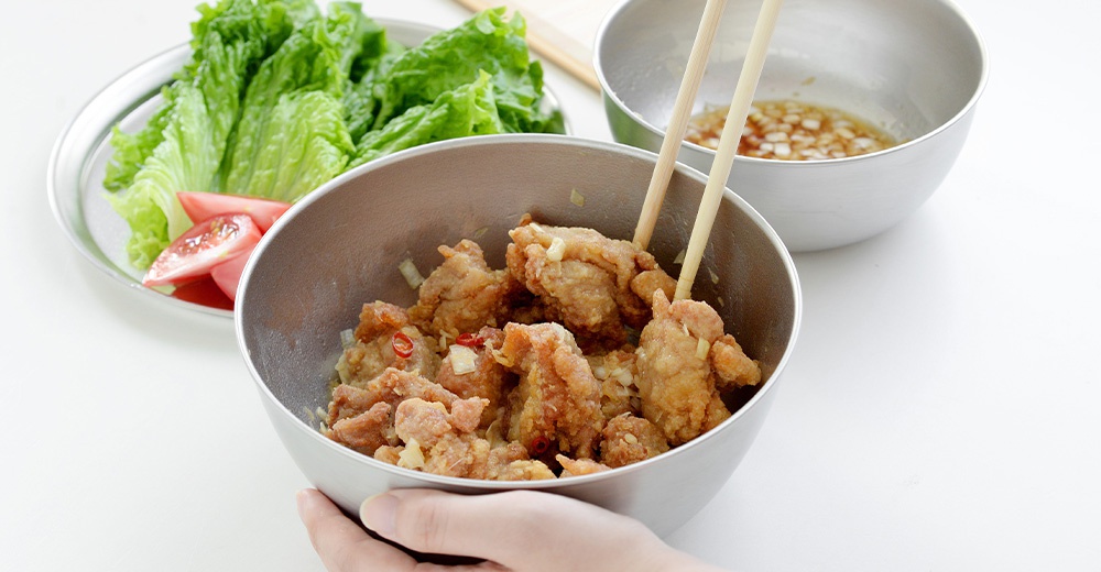 炸雞塊盛裝在圓形鋼碗中，用筷子夾起，碗旁有生菜和番茄切片，另一鋼碗中裝有醬汁。