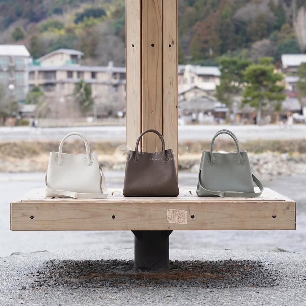三款京都職人手作兩用手提托特包，分別為米白色、咖啡色和灰綠色，皆為皮革材質，設計簡約，配有提把與可拆卸肩背帶，置於木製長椅上。