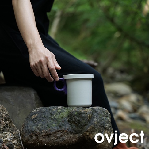 一個象牙白琺瑯馬克杯，配有紫色提把，置於岩石上，背景為模糊的綠色樹林。