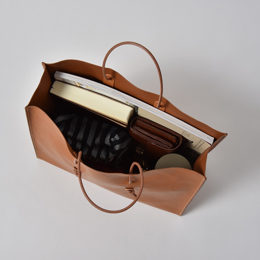 職人手作皮革提袋，淺棕色，光滑皮革材質，設計簡潔，可容納書籍、錢包、條紋織物等物品。