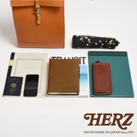 淺棕色皮革製成的紙袋風後背包，搭配黃銅金屬扣環。旁邊擺放著一本深棕色皮革筆記本、一個深棕色皮革長夾、一本雜誌、一本護照、一支手機及一把印有彩色圓點圖案的黑色摺疊傘。