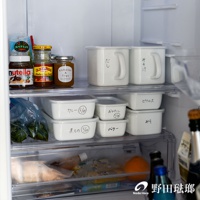 兩個白色帶把手的方形儲物罐，其中一個罐子寫著「だし」，另一個寫著「みそ汁」。下方層架有數個白色長方形儲物罐，其中標示著「カレー (5/27)」、「おひたし (5/28)」、「煮もの (5/28)」、「バター」和「ピクルス」等字樣。