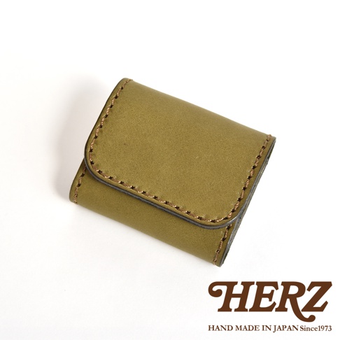 一個橄欖綠色的日本HERZ職人手作皮革雙折收納零錢包，表面呈現細膩的皮革質感，邊緣有明顯的縫線細節，設計簡潔且外型小巧，右下角印有HERZ品牌商標與產地說明。