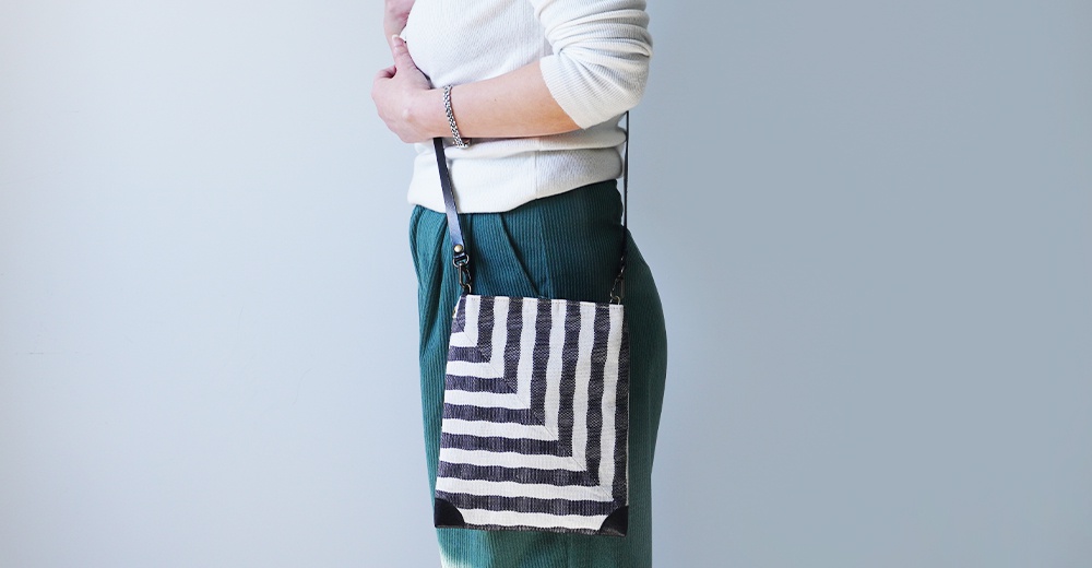 BAN INOUE 斑馬條紋麻布散步斜背包，由左側視角拍攝，一位女性穿著白色長袖上衣與綠色燈芯絨長褲，斜背著一個黑白條紋的麻布方包，包包底部有黑色皮革裝飾。