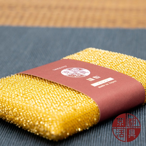 金黃色不沾鍋專用菜瓜布，表面佈滿細小顆粒，材質為纖維網狀。產品以紅色紙帶綑綁，紙帶上印有白色文字與圖案，包括「SINCE 1717」、「錦」、「製造」、「スポン」、「made in Japan」以及一個圓形紅色標誌。
