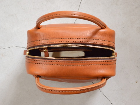 職人手作皮革Organ方形托特包，皮革呈現溫潤的橘棕色，包身線條俐落，以車縫線勾勒出方形輪廓，頂部設有雙提把，拉鍊開口露出部分淺色內襯及一本小書。
