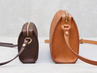 兩個皮革拉鍊單肩包，一個深棕色，一個棕褐色，皆為圓潤的造型，配有金色拉鍊和皮革背帶。