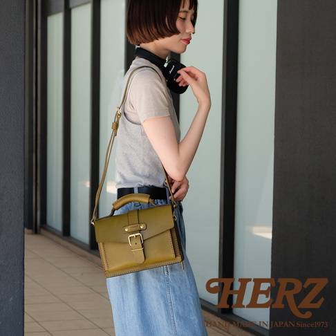 一位女性側身站立，肩背一個橄欖綠色皮革迷你單肩包。包包為方形，配有頂部提把、可調式長斜揹帶，以及一個以皮帶和黃銅釦固定的翻蓋。包包表面有精細的縫線，展現職人手作質感。