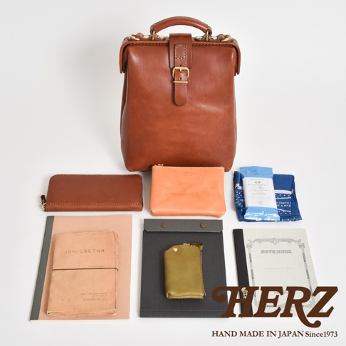 職人手作皮革迷你軟皮杜勒斯包，搭配同色系長夾、橘色化妝包、綠色零錢包、兩本筆記本、藍色印花手帕及一包濕紙巾，展現精緻的皮革工藝。