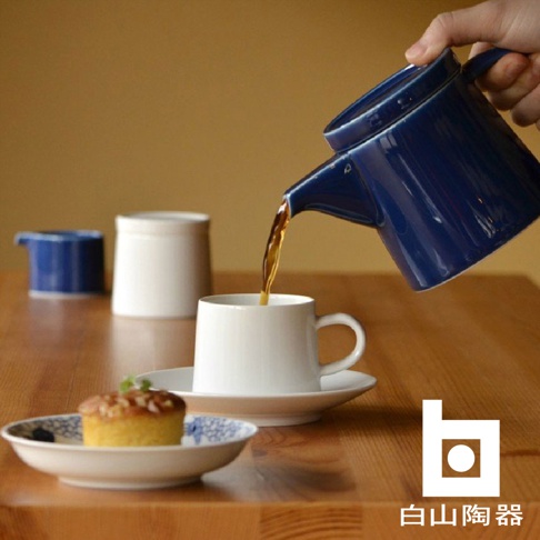 藍色陶瓷M型壺傾倒茶水至白色陶瓷杯中，背景有白色和藍色陶瓷罐，以及裝有小蛋糕的盤子。