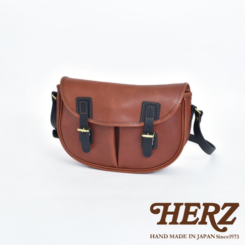 職人手作皮革傳統釣魚包，採用棕色皮革，配有黑色皮質翻蓋、雙皮帶扣和金色金屬扣。包身呈現半月形，帶有肩帶。