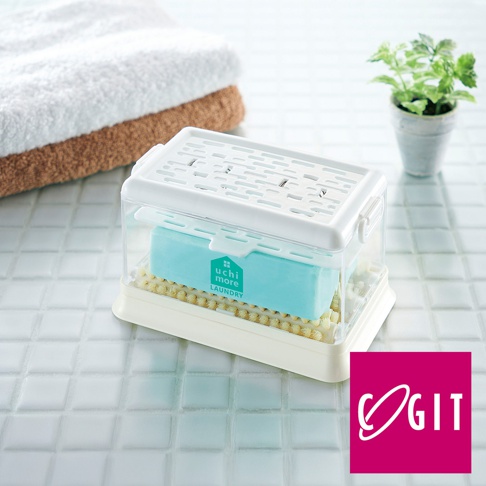 白色塑膠製的多功能免沾手泡沫洗衣皂盒，盒蓋上有通風孔。盒內有一個淺藍色的洗衣皂，置於黃色的刷毛底座上。背景是堆疊的毛巾和綠色植物。