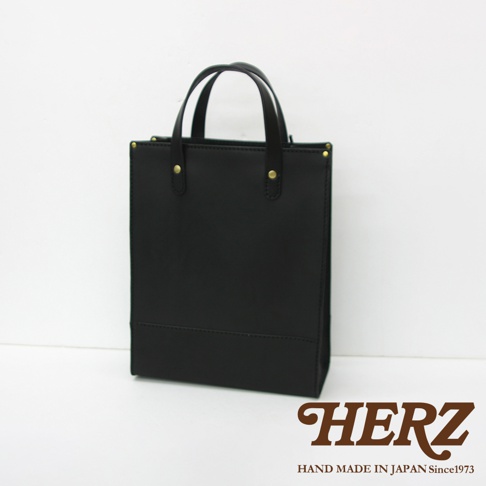 黑色皮革直式托特包，簡約設計，頂部有提把，兩側有金色卯釘裝飾，底部有橫向車縫線。