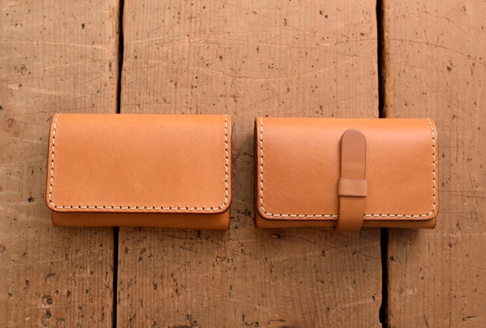 兩個淺棕色皮革名片夾，皆為長方形，附有手工縫線。左側名片夾為素面，右側名片夾則有一條皮革帶子，可固定夾身。