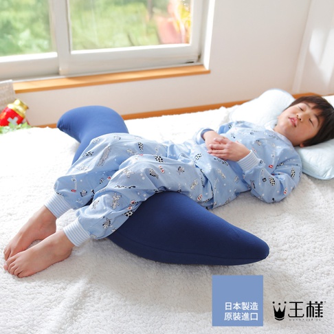深藍色「王樣の兒童抱枕」呈彎月形，柔軟布料包裹，一位穿著藍色卡通圖案睡衣的男孩側臥在其上，頭枕著淺藍色枕頭，整體畫面呈現舒適的居家氛圍。