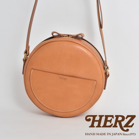 職人手作皮革小圓包，天然棕色，光滑皮革材質，圓形設計，正面有半月形口袋，拉鍊開合，附有可調式皮革背帶。