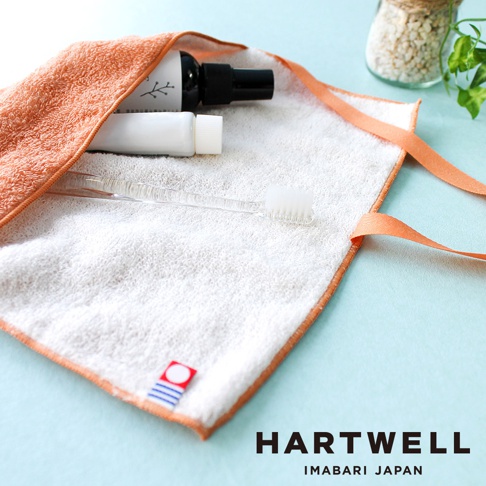 橙色毛巾收納袋內含一瓶黑色噴霧狀物、一條白色牙膏軟管，以及一支透明牙刷。收納袋邊緣為橙色滾邊，並配有兩條橙色細繩。