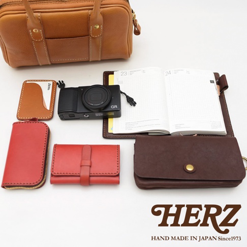 HERZ 品牌皮革製品，包含一個棕色迷你波士頓包，一個黑色相機，一本日誌本，一個紅色零錢包，一個紅色卡夾，以及一個深棕色皮夾。