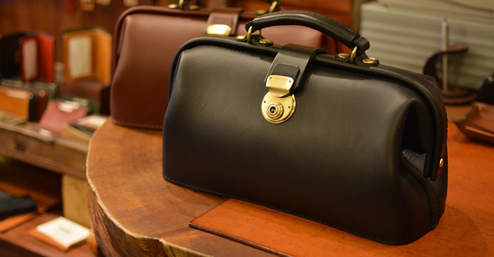 黑色皮革迷你醫生包，配有金色旋轉鎖扣和皮革手柄，另一款棕色皮革醫生包在後方。