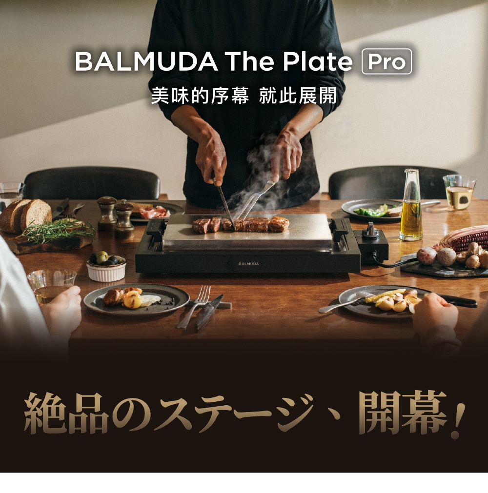 黑色 BALMUDA The Plate Pro 極致電烤盤，帶有銀色烤盤，上面正在烹調牛排。周圍擺放著餐盤、麵包、蔬菜和調味料。