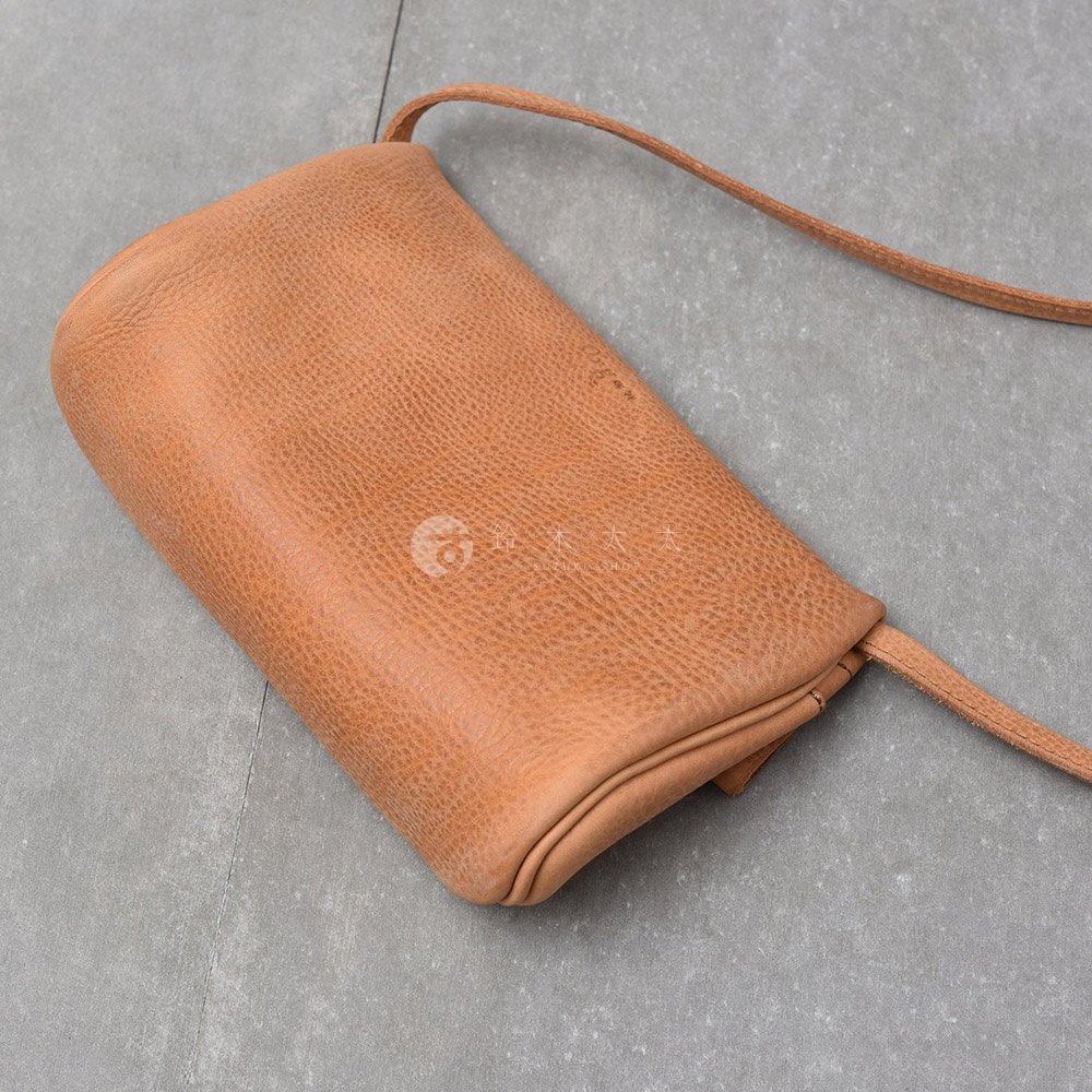 一個淺棕色的皮革斜背包，包身呈現柔軟的弧度，表面有自然的紋理，附有細長的同色系背帶。