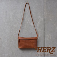 一個棕褐色的皮革斜背包，正面有拉鍊，掛在灰色紋理的牆壁上，下方有HERZ品牌標誌和手作字樣。