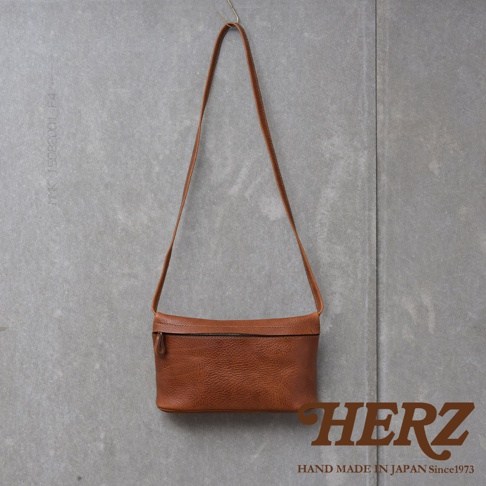 一個棕褐色的皮革斜背包，正面有拉鍊，掛在灰色紋理的牆壁上，下方有HERZ品牌標誌和手作字樣。