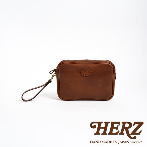 棕色皮革手提包，附有可拆卸的皮製腕帶和金色扣環。包包正面中央有一個壓印的圓形標誌，標有「HERZ」字樣。
