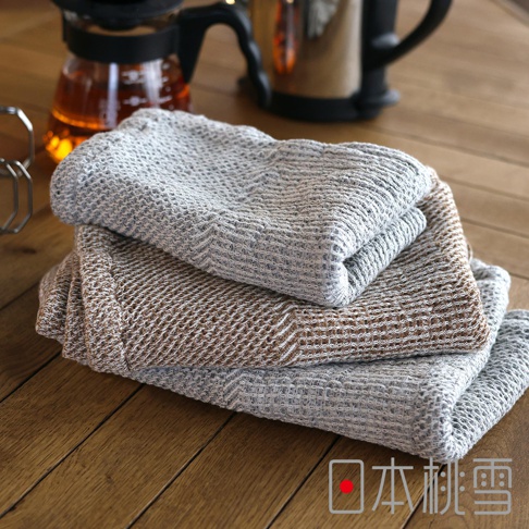 日本桃雪SEK抗菌防臭檜木鬆餅紋毛巾，疊放在木桌上，上方為灰色，中間為咖啡色，下方為灰色，毛巾呈現鬆餅紋理，背景有玻璃茶壺。