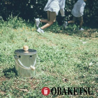 OBAKETSU 品牌標誌旁是手工製的金屬蚊香桶，桶蓋上有木製圓球裝飾，桶身側面也有木製把手，背景是模糊的綠色草地和奔跑中的兩個人。