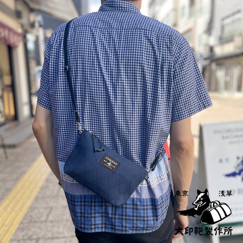 藍色刺子織隨身斜背包，方形格紋縫線，配有黑色斜揹帶和金屬扣環，包上貼有紅色與白色標籤，由一位身穿藍白格紋襯衫的男士斜揹於身。