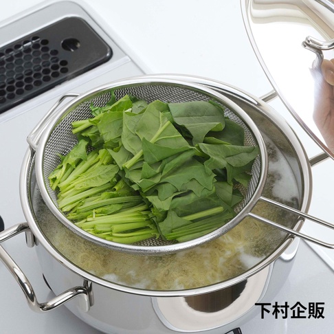不鏽鋼料理濾網，呈現銀色金屬光澤，網孔細密，裝有大量新鮮的綠葉蔬菜，置於熱水中冒著蒸氣的鍋子內。