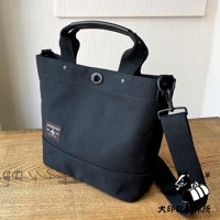 黑色帆布材質的兩用托特包，設有黑色皮革提把、可拆卸調節的帆布斜背帶，以及正面紅色標籤，標籤上印有日文圖案和文字。