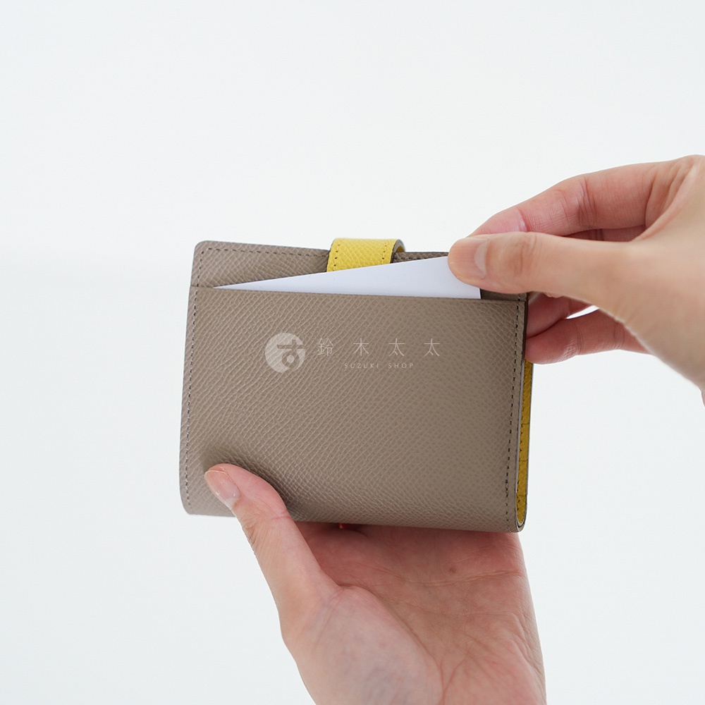 一只手拿著一個灰色皮革短夾，夾內露出部分白色卡片，另一隻手正在調整卡片。短夾內側有黃色裝飾，左側印有日文「鈴木太太 SUZUKI SHOP」字樣。