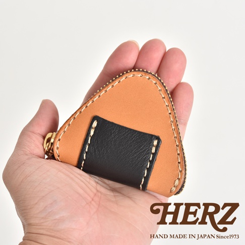 手掌上捧著一個日本HERZ手工製作的皮革零錢包，外觀呈現可愛的三角飯糰造型，主體為淺棕色皮革，搭配正中央的黑色皮革拼接裝飾，邊緣縫線細緻，並鑲有拉鍊，右下角印有品牌名稱與創立年份資訊。
