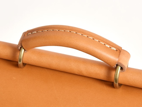 職人手作皮革屋瓦風迷你後背包 (R-6) 的特寫，展示了淺棕色皮革手柄，帶有精緻的縫線和黃銅色金屬環，連接到圓柱形的皮革部分。