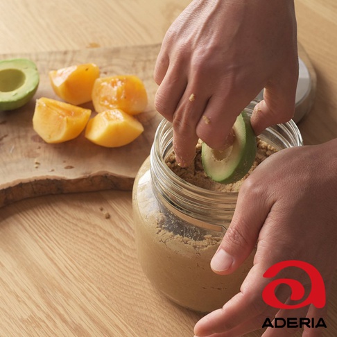 一隻手將切片的酪梨放入裝有棕色粉末的透明玻璃罐中，罐旁有切塊的黃色水果。