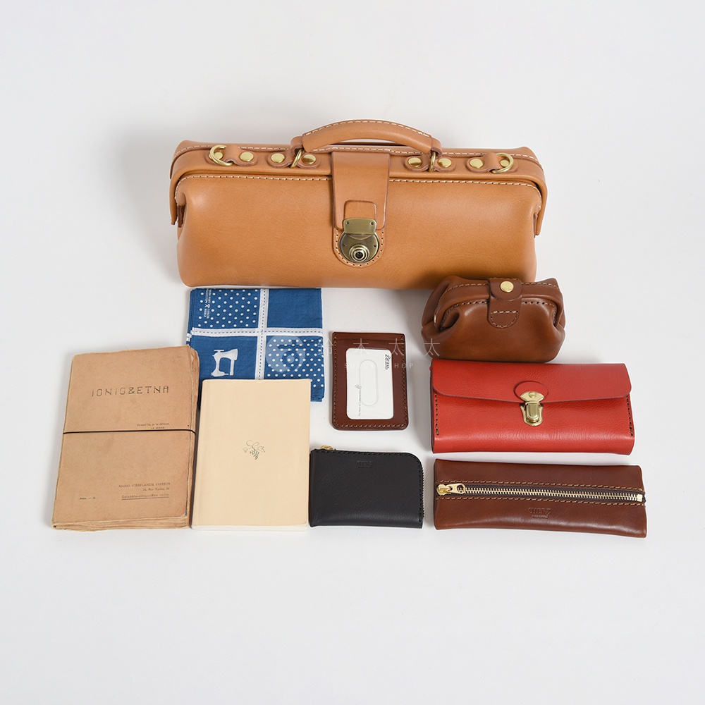 圖中有一個淺棕色皮革製作的波士頓包，下方擺放著一個藍色方巾、一個棕色皮革零錢包、一張紅色的皮夾、一個深棕色長夾、一個淺棕色小筆記本，以及一個印有 IONIO&ETNA 文字的檔案夾。