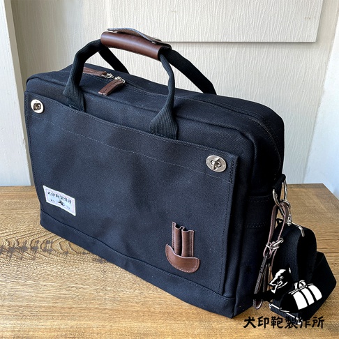 黑色純棉帆布商務公事包，B4尺寸，設有皮革提把、拉鍊開合、側邊旋轉鎖扣及可拆卸肩帶。包面上方有品牌標籤，下方裝飾有皮革飾件。