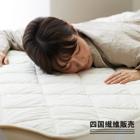 一張雙人加大尺寸的米白色羊毛床墊，表面有規則的絎縫格紋，一位女子正舒適地趴臥在床墊上。