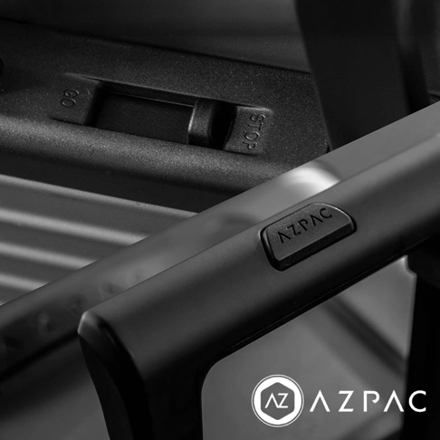 黑白攝影視角呈現 AZPAC 行李箱細節，特寫鏡頭展示了行李箱拉桿手把上印有 AZPAC 品牌壓紋，後方隱約可見帶有 GO 與 STOP 標示的煞車裝置，整體色調為深灰色，風格簡約現代。