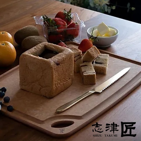 金屬材質的麵包刀，帶有鋸齒狀刀刃，刀柄為金色，置於淺棕色木質砧板上，旁邊是方形吐司、草莓、藍莓、橘子、奇異果及一碗奶油。