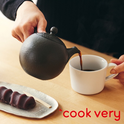 一只手正從黑色常滑燒咖啡急須茶壺中，向一個白色馬克杯倒咖啡。茶壺帶有紋理的表面和圓潤的壺身。一旁銀色盤子上有紅豆麻糬和一根木籤。