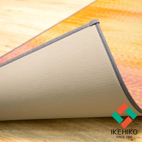 藺草榻榻米瑜珈墊，表面為天然藺草編織，帶有橙色與粉色漸層的抽象圖案。墊子側面邊緣包覆灰色布料，底部為淺米色止滑材質，平鋪於木紋地板上。