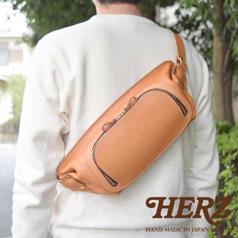 日本HERZ職人手作皮革旅行斜背包，展示在穿著白色毛衣的人背上，特寫包包的棕褐色皮革材質與拉鍊細節