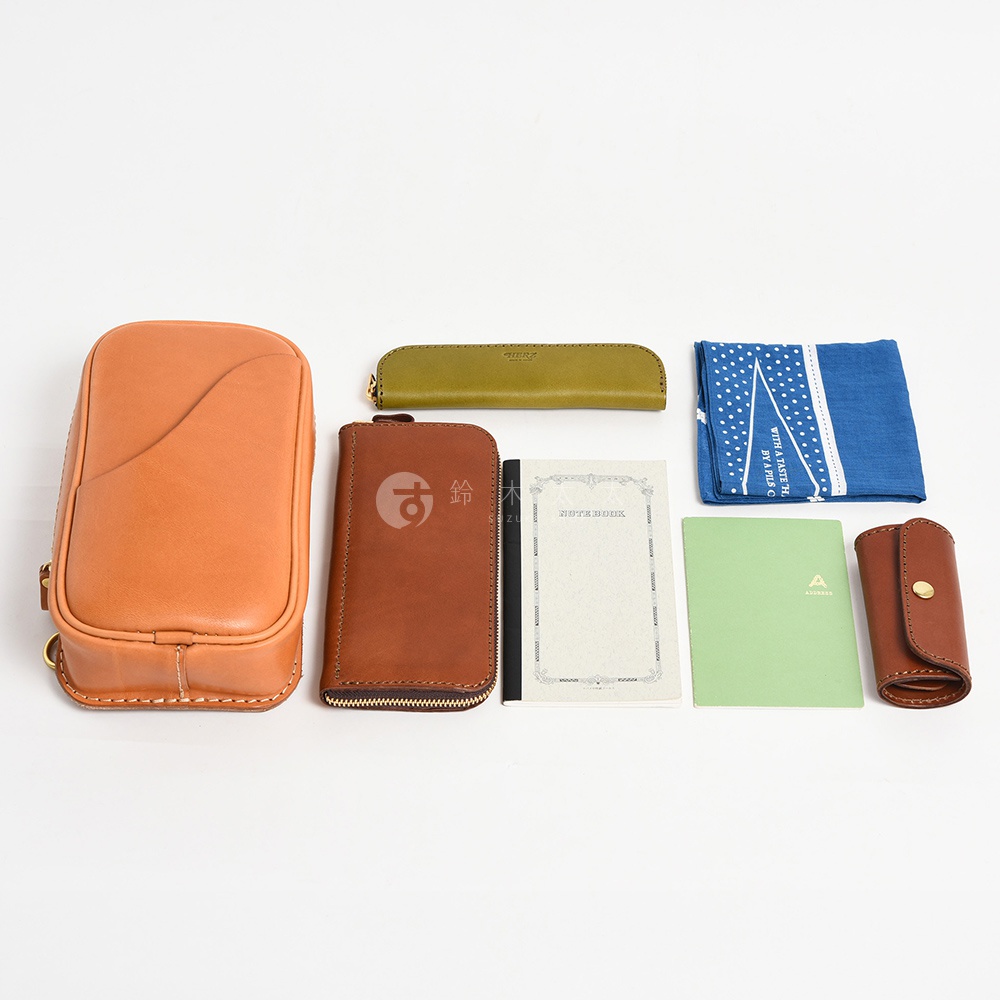 日本HERZ職人手作皮革兩用直立型斜背包，包含一個淺棕色皮革斜背包、一個深棕色皮革長夾、一支綠色皮革筆套、一本白色筆記本、一本綠色筆記本、一個藍色圖案手帕和一個咖啡色皮革鑰匙包。