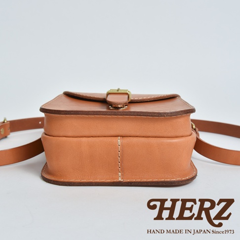這是一款日本HERZ手工製作的Latigo硬皮革迷你單肩包，側面視角展示其挺立的包體結構與厚實皮革質感，配有金屬扣環與可調式背帶，整體設計簡約經典並印有HERZ品牌標誌。