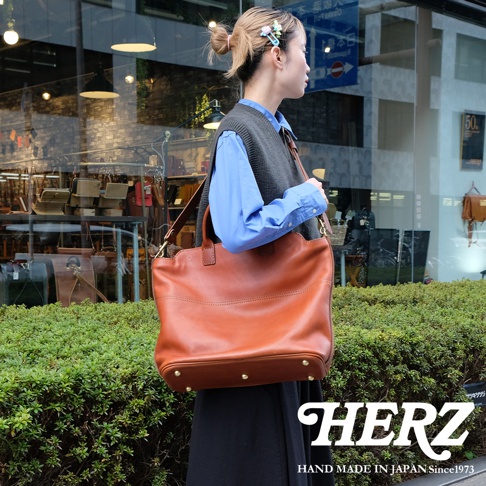 棕色皮革大容量托特包，配有可拆卸肩帶，包包底部有四個金屬腳釘，整體呈現簡約大方的設計風格，展現職人手作質感。