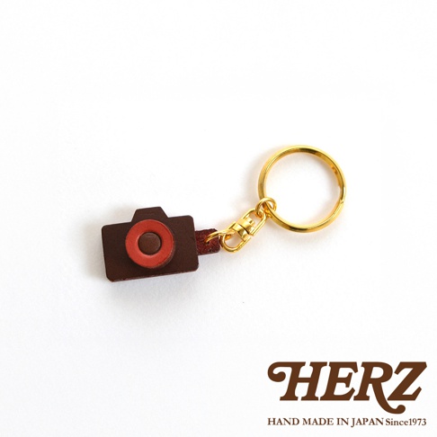 職人手作皮革相機造型鑰匙圈，深棕色皮革製成，搭配紅色鏡頭裝飾，附有金色金屬掛環及旋轉扣。